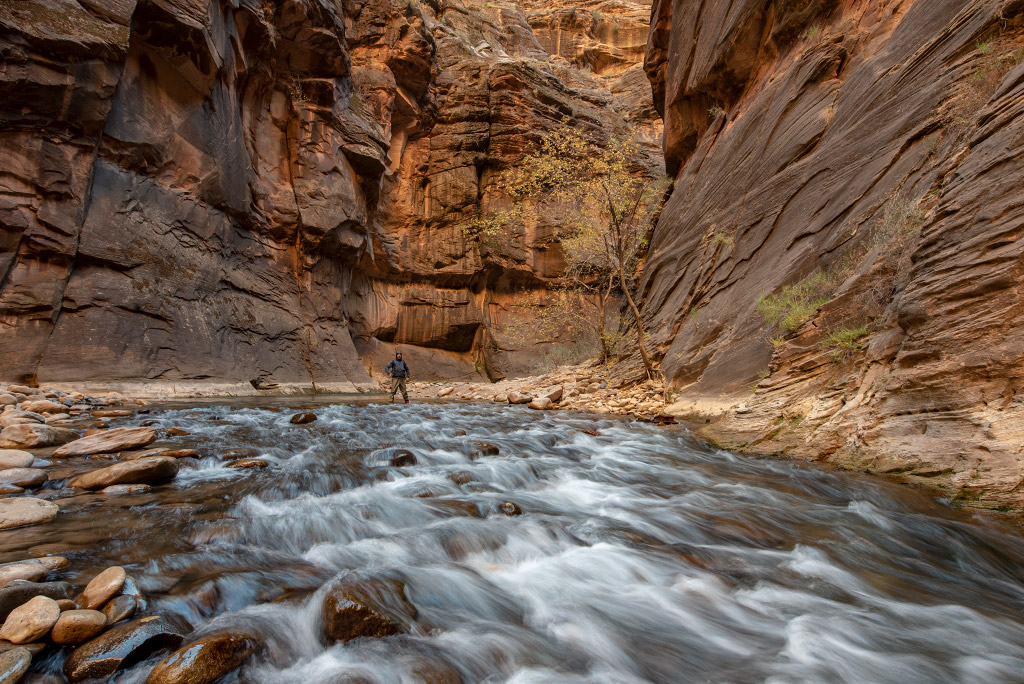 Chris is tanding in the river among tall walls during The Narrows hike in Zion National Park.