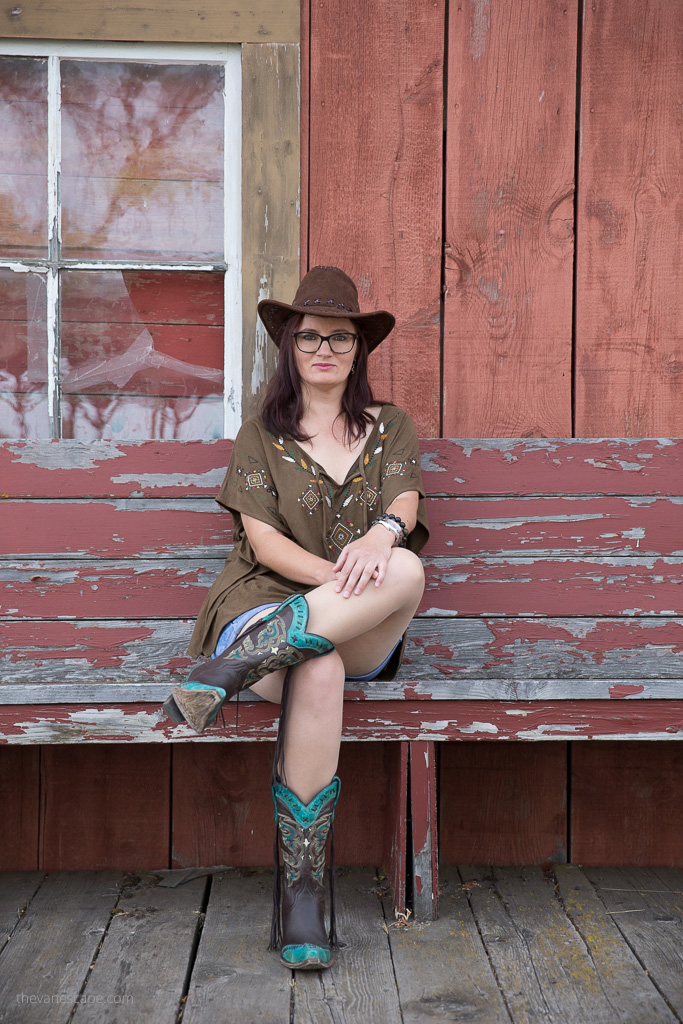 Agnes Stabinska, the author, is wearing brown and blue cowboy boots, a denim skirt, a brown blouse and a large brown cowboy hat. She sits on a wooden bench in Shaniko Ghost Town.