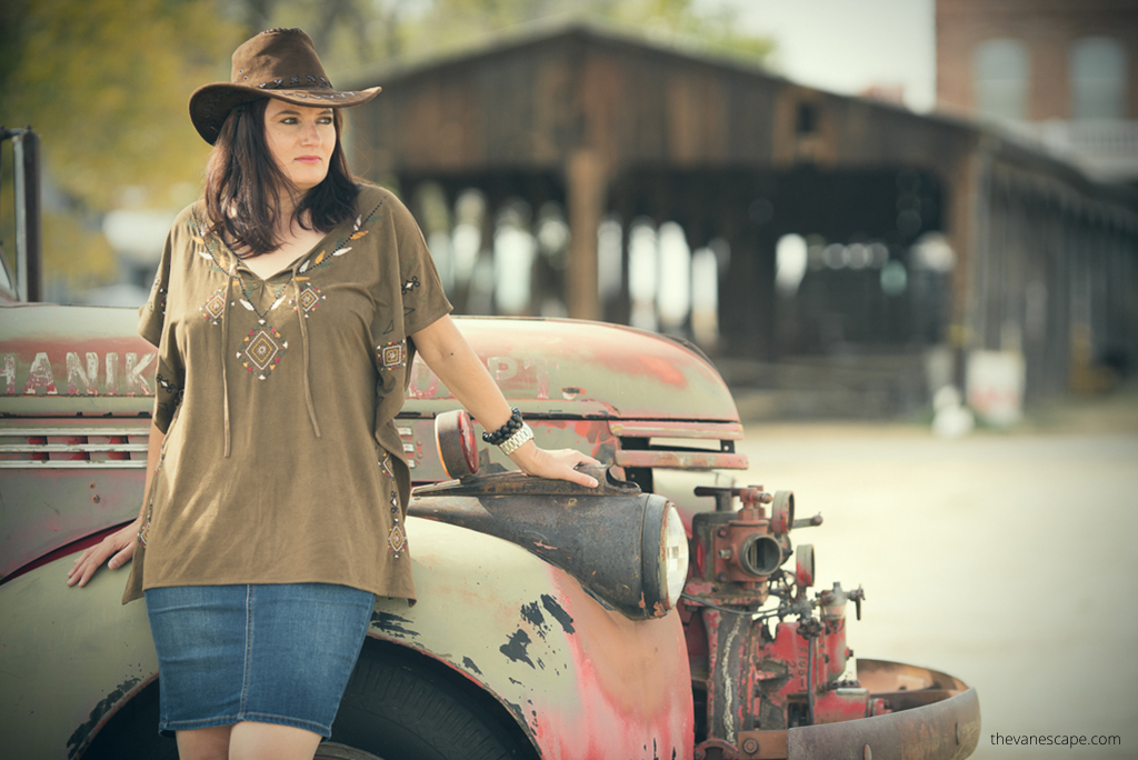 Agnes Stabinska, the author, wears cowboy hat in Shaniko Ghost Town.  She is next to old car and with old wooden buildlings in the backdrop.