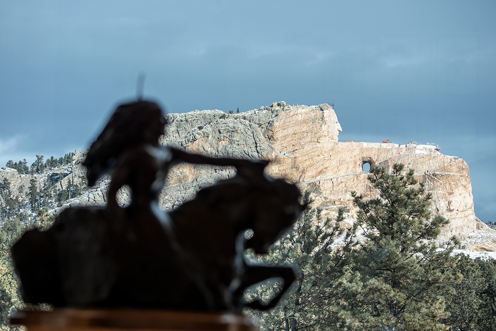 In the foreground, a bronze model of Crazy Horse stands proudly on a platform inside the museum, while in the background, the massive mountain carving continues to take shape.