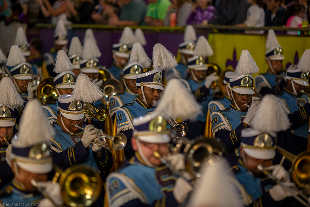 A brass band roams the streets of New Orleans, they play trumpets and trombones, wearing caps with plumes on their heads.