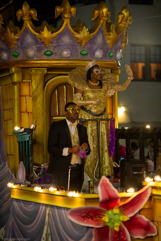 A woman in a gold dress, gold crown, and gold face mask waves to the audience from a platform in New Orleans.